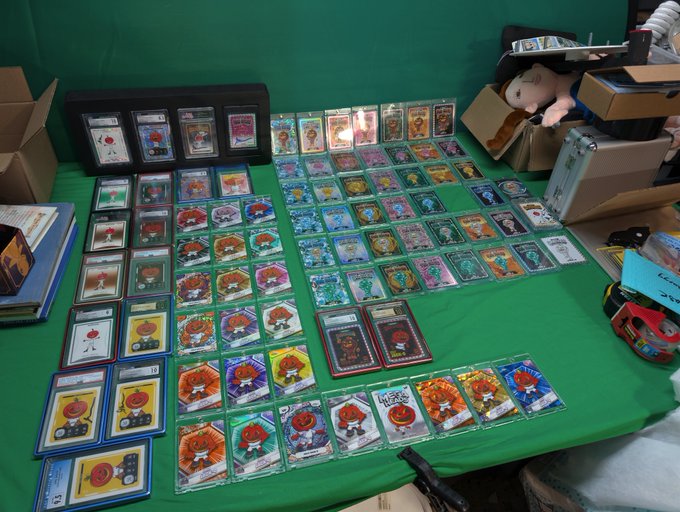 A green tabletop is covered with numerous colorful trading cards displayed in protective sleeves, alongside binders, loose cards, and boxes of merchandise.