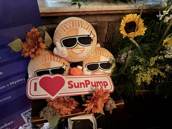 A 'I ❤️ SunPump' sign featuring three cartoon sunflower characters wearing sunglasses and giving thumbs up, displayed alongside decorative flowers.