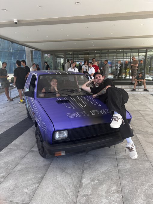 Two people pose on a purple Volkswagen Golf with 'SOLANA' written across the hood, parked outside a building with bystanders watching.