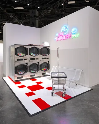 An art installation titled 'The Laundromat' displaying washing machines, a shopping cart, and neon signage on a red and white checkered floor.