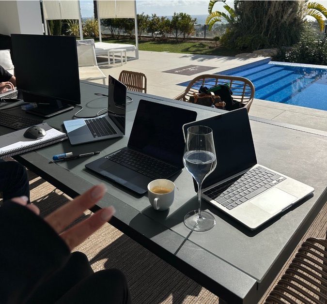 An outdoor work setup by a swimming pool with multiple laptops, a monitor, a wine glass, and an espresso cup on a table, while a person in the foreground makes a peace sign.