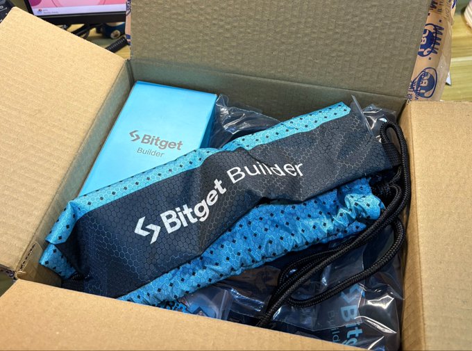 The image shows a cardboard box containing a blue and black backpack, along with other items such as a pair of sunglasses and a phone case. The text "received an unexpected care package from @BitgetBuilders" suggests that someone has sent these items as a surprise gift or gesture of support for the Bitget Builders community.