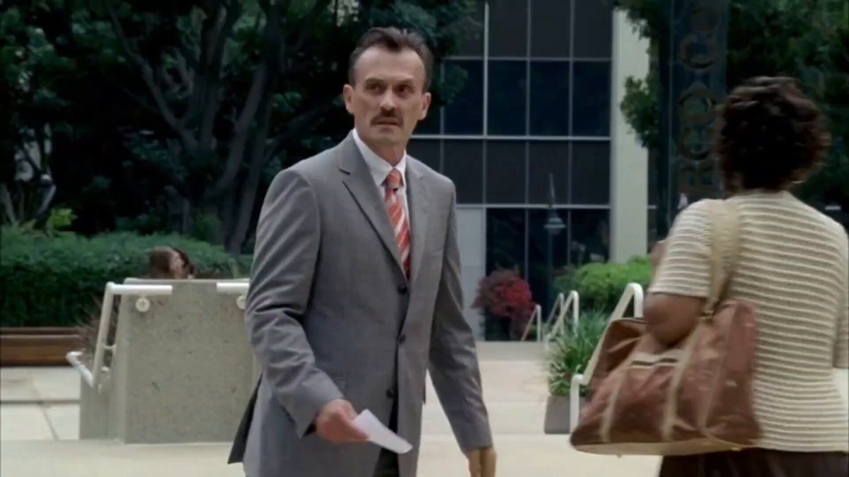The image shows two people standing outside of an office building. One person is wearing a suit and tie, while the other has a handbag over their shoulder. They appear to be engaged in conversation or observing something happening inside the building. The scene suggests that they might be discussing work-related matters or attending a business event together.