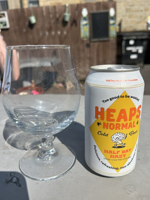 A beer glass and a can of 'Heaps Normal Half Day Hazy' sitting on a concrete outdoor table, with the can describing the beverage as 'Too good to be wasted' and 'Cold Beer' that is 'Delicious, refreshing, non-alcoholic' at 3.7% alcohol.