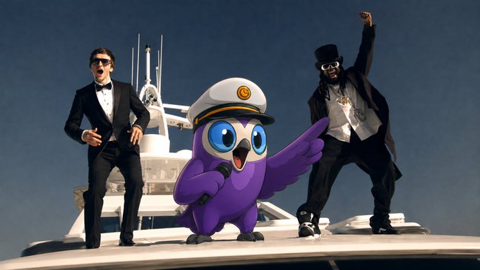 A purple cartoon owl mascot wearing a white captain's hat points to the right while standing on a yacht between two men in formal black attire who are posing enthusiastically.