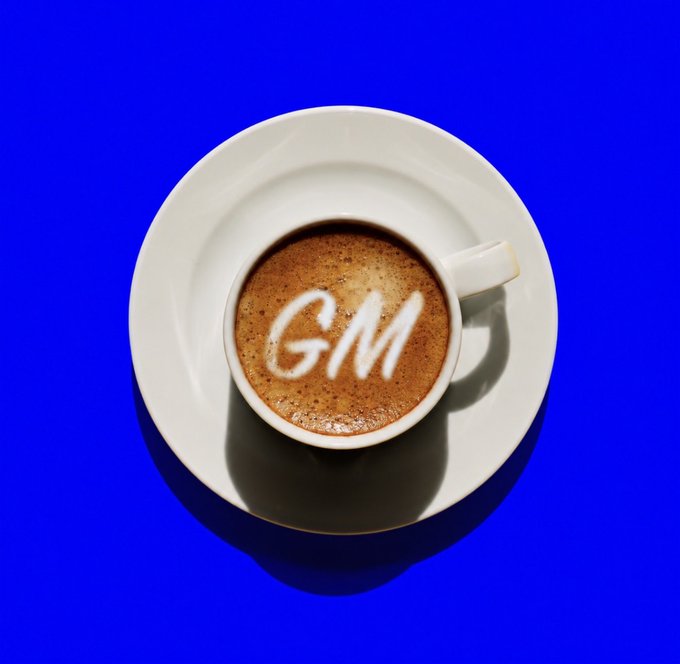 A white cup of coffee on a white saucer against a blue background, with "GM" written in milk foam on the surface.