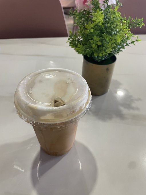 An iced coffee in a clear plastic cup with a lid sits on a white marble table next to a small potted green plant.