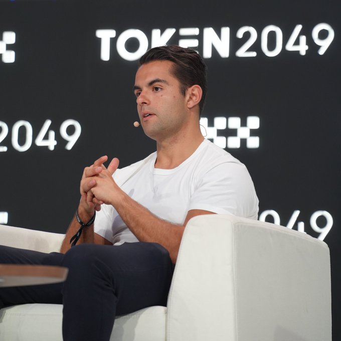 A man in a white t-shirt speaks on stage at the TOKEN2049 conference, seated in a white chair with his hands clasped.