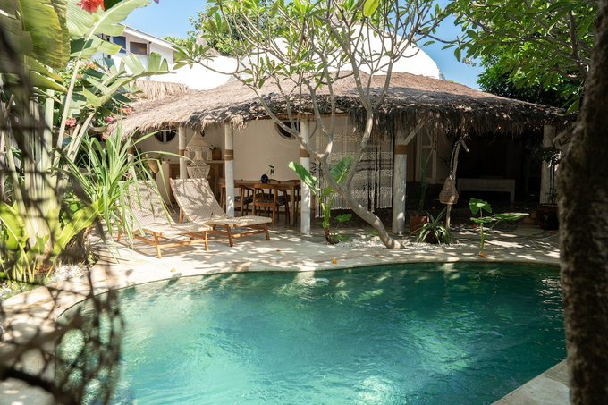 A serene tropical villa features a thatched-roof open-air lounge beside a turquoise swimming pool, surrounded by lush frangipani trees and lounge chairs. The tranquil setting evokes the relaxed, exploratory spirit of the accompanying post, inviting viewers to imagine discovering such hidden paradises.