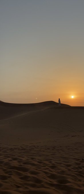 A silhouette of a person standing on a sand dune as the sun sets, creating a warm orange glow against the darkening sky.