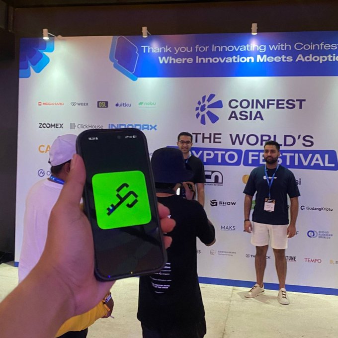 A person holds up a smartphone displaying the Syndicate (green key icon) app logo while photographing attendees at Coinfest Asia, the world's largest crypto festival, capturing the community's presence at major crypto events.