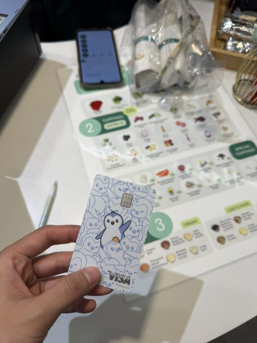 A hand holds up a Pudgy Penguins-themed Visa debit card featuring a cartoon penguin and "VISA" text, set against a backdrop of food menus and a smartphone at what appears to be a restaurant or café counter. The image ties to the caption by showing the custom KAST crypto card being used casually for everyday purchases like a salad.