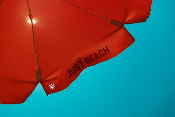 A red umbrella bearing the text "JUST BEACH" against a teal sky. The image's leisurely "just beach" sentiment creates an ironic contrast with the post's emphasis on the often-overlooked energy demands of DeFi transactions, suggesting that what appears to be a simple, relaxing activity can carry hidden resource costs.