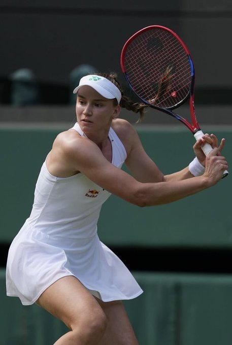 Tennis player Donna Vekić in a white outfit and white visor hits a two-handed backhand with a red and blue racket during a match.
