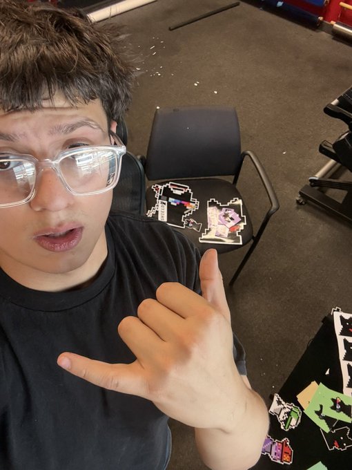 A young man with glasses taking a selfie while pointing at pixel-art stickers on a black chair behind him.