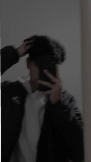 A person wearing a black and white North Face jacket takes a mirror selfie while holding a phone, with one hand in their hair, appearing slightly blurry and dark.