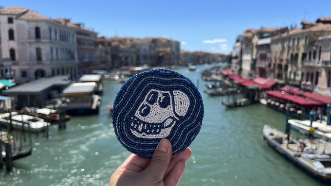 The image shows a hand holding up a blue and white coaster featuring a skull design, likely made of beads or yarn. The coaster appears to be placed over a body of water, possibly a canal or river, which adds an interesting visual element to the scene. The caption above the image reads "GM fam Enjoyed a day trip to Venice yesterday Always travelling with my ApeCoin coaster from Rich Class Decor". This suggests that the person holding up the coaster is likely on a trip to Venice and has brought along their unique coaster as a souvenir or decoration for their accommodations.
