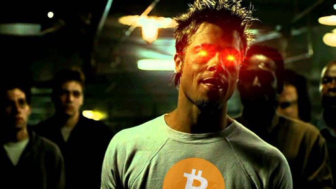 A man with glowing red eyes stands in a dimly lit industrial setting wearing a grey sweatshirt with an orange Bitcoin '₿' logo, with several other people standing behind him.