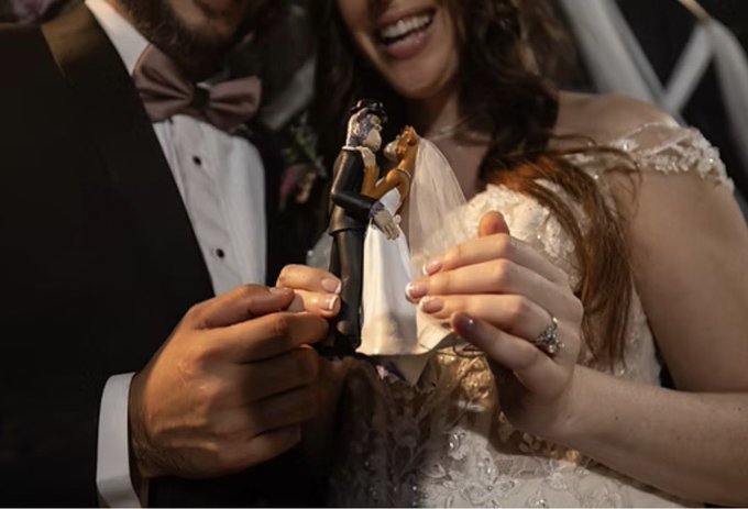 A newlywed couple holds a wedding cake topper of a bride and groom, with the bride's ring visible on her hand.