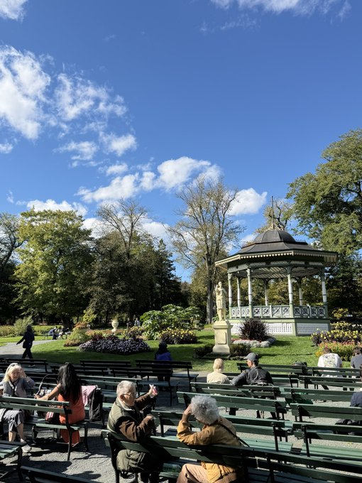 People sit on green benches in a sunny park enjoying a clear day, with a white ornate bandstand, lush trees, and flower beds in the background.