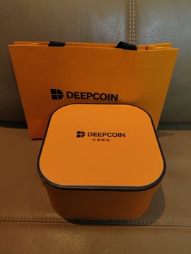 A Deepcoin-branded orange shopping bag sits behind a square orange tin box, both displaying the "DEEPCOIN" logo with Chinese characters underneath. The packaging matches the post celebrating a Mid-Autumn Festival gift box from Deepcoin, which contained an aromatic diffuser set.