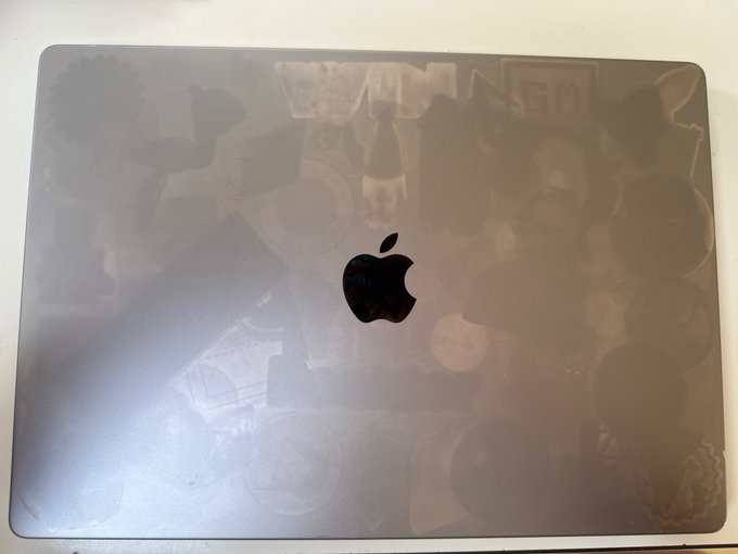 A closed silver Apple MacBook laptop sits on a surface, with reflections of stickers visible on its lid.