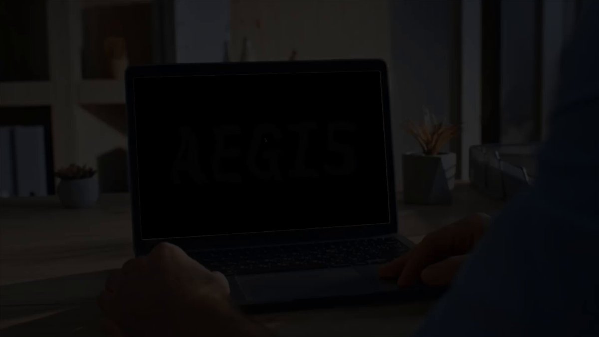 The image shows a person sitting at a desk with a laptop computer open in front of them, displaying the word "AEGIS" on its screen. The background features a windowed office space with various items such as potted plants, books, and a vase.
