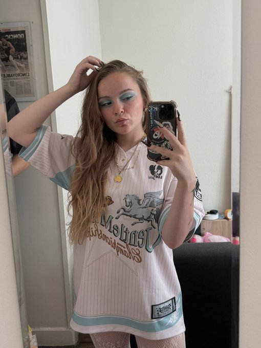 A mirror selfie of a young woman with long brown hair and blue eyeshadow, wearing a striped blue and white jersey with 'Los Leones' and 'Stadium' branding, holding a phone with a decorated case.