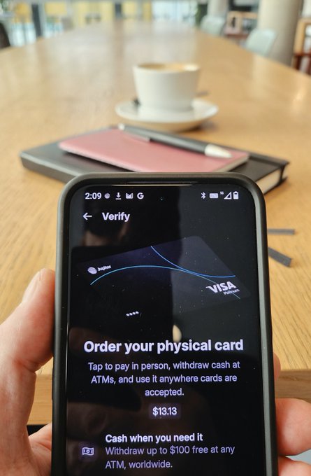 A hand holds a phone displaying a "Verify" screen with a Visa virtual card from Sugoi text and a $13.13 balance, with the prompt "Order your physical card" visible on a café table.
