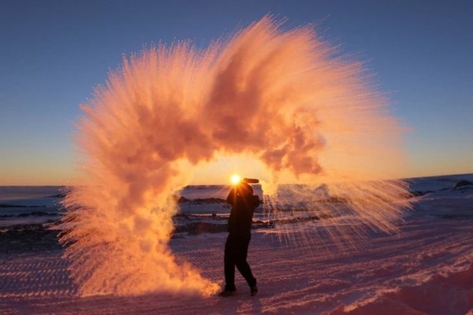 A silhouetted figure throws a bucket of water into the air, creating a stunning frozen archway of orange-glowing water droplets against a sunset sky in a snowy landscape.