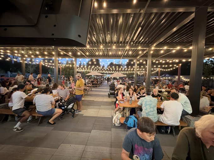 A crowded outdoor patio or beer garden at dusk, filled with people sitting at long wooden tables under a covered structure with string lights.