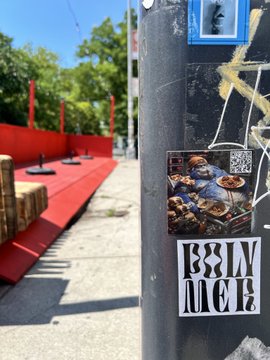 Street photography shot showing a metal pole with stickers, including a "BIM HEIR" decal, with a red platform or bench and trees visible in the background on a sunny day.