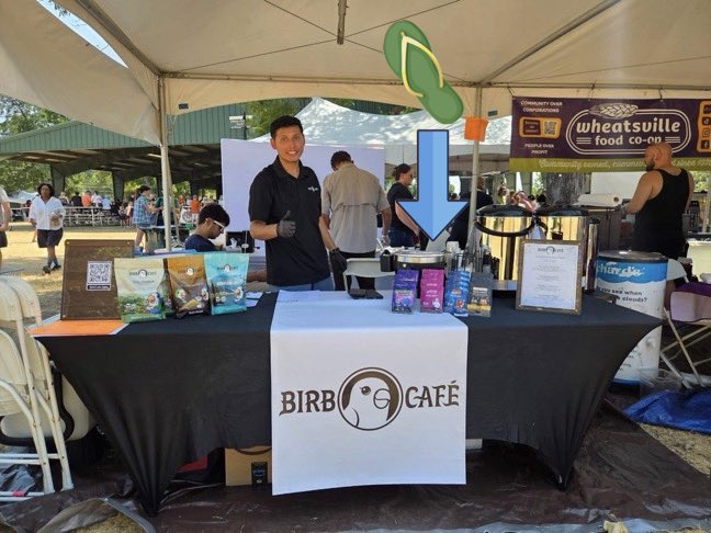 A vendor booth at an outdoor market for 'BIRDBOY CAFÉ', with a smiling man in a black polo standing behind a table displaying packaged snacks, with a blue arrow pointing to the products.