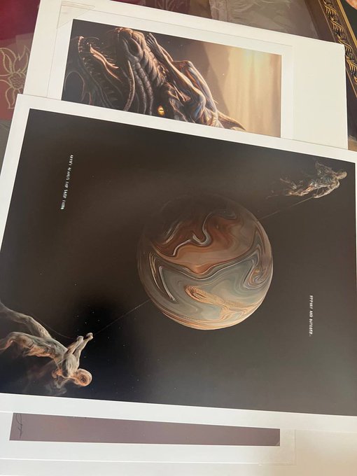 Two printed art pages overlapping on a surface, depicting space scenes with planets and figures, including one showing a swirled orange and white gas giant planet flanked by two floating humanoid figures.