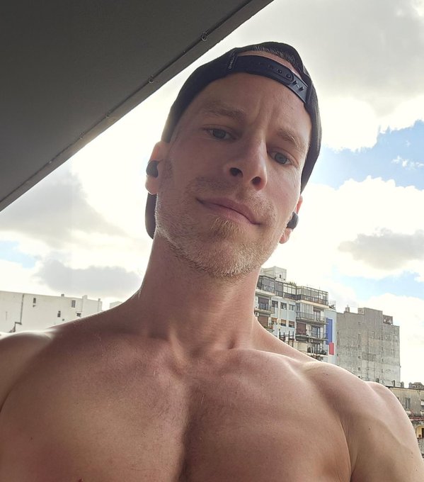 A shirtless selfie of a young man wearing a backward black cap and earbuds, photographed outdoors against a backdrop of buildings and a cloudy sky.