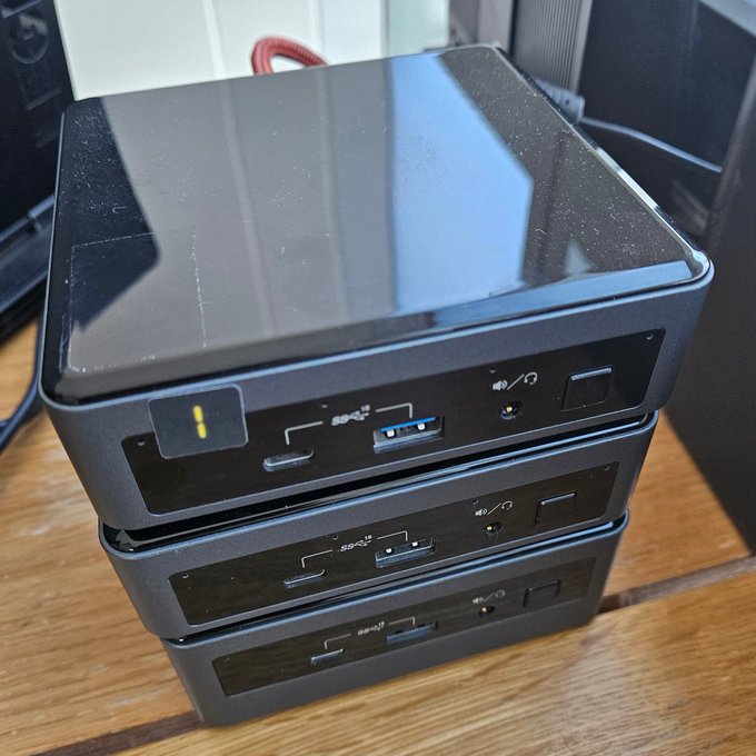 A stack of three black mini PC units with USB ports sits on a wooden desk, showing wear and scratches on the glossy top surface.