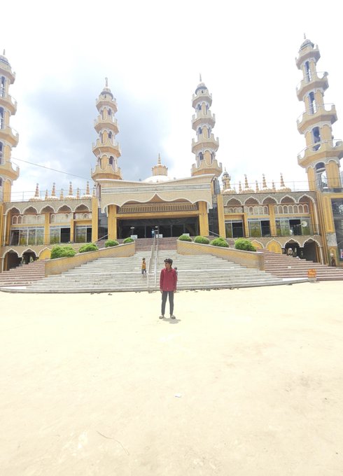 A wide-angle photograph of a person standing on a sandy plaza in front of a grand, yellow mosque with multiple tall minarets and a central dome.