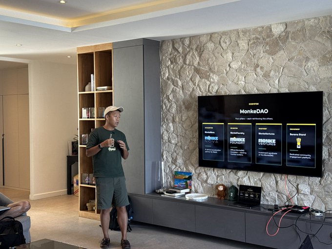 A man in a cap and shorts stands in a modern living room in front of a TV displaying "MonkeDAO" alongside various project categories including MonkeFund, MonkeMarketplace, and Banana Stand. The image visually supports the post's theme of networking and community-building, capturing a casual in-person connection moment within a comfortable home setting that reflects the personal, organic nature of the Web3 meetups described.