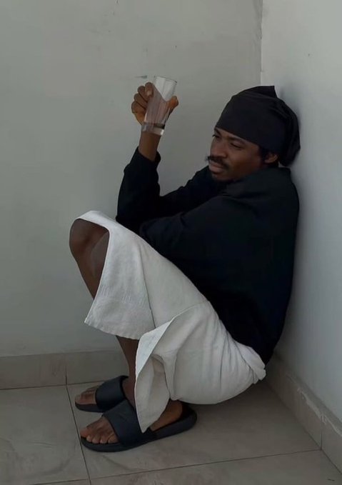A person wearing a black hoodie, white shorts, and a black head wrap sits on the floor against a white wall, holding a clear glass of water. They are wearing black slide sandals and looking off to the side.