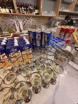 A cluttered bar counter covered with numerous empty drinking glasses, blue 'Asahi' beer cartons, and tall beer cans, with bottles lined up on shelves in the background.