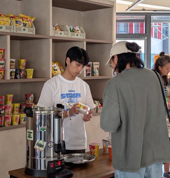 A young man wearing a "Stark" branded t-shirt pours instant noodles from a pot into a bowl for a customer at StarkMart Seoul, with shelves of snack products visible in the background. The image captures the "hot noodles, hotter tech" tagline in action—blending convenience-store food culture with Stark's brand presence at the Seoul pop-up.