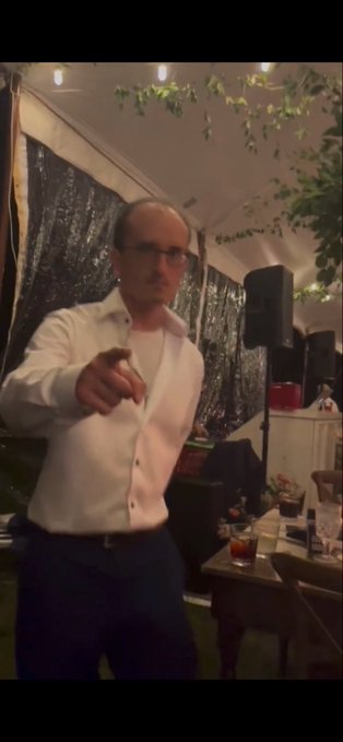 A man in a white button-down shirt and glasses points toward the camera while standing under a tent decorated with string lights and greenery, likely at an evening event. The image humorously illustrates the joke about giving @CryptoGarga a Sophie lookalike hairstyle to pump the floor price of the "Sophie" ape NFT collection.
