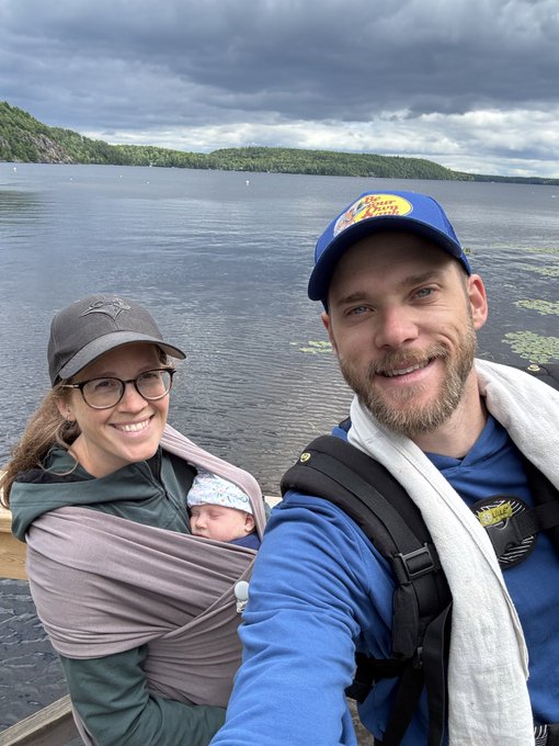 A selfie of a family of three—a man in a Bass Pro Shops cap, a woman in glasses with a baseball cap, and a sleeping baby in a carrier—by a lake with forested hills in the background.