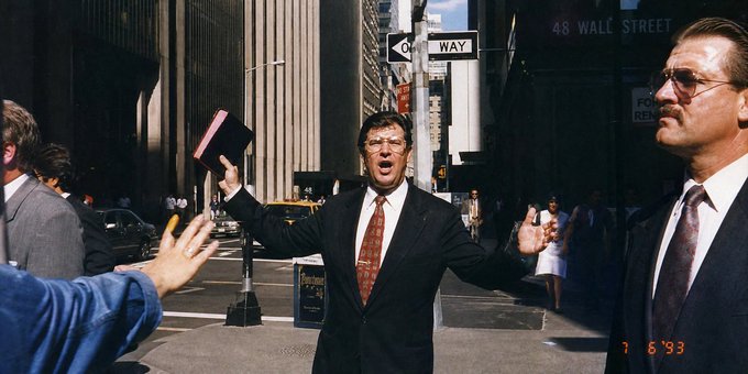 A photograph dated "7 6'93" showing a man in a dark suit and glasses holding up a book on Wall Street, with a sign reading "48 WALL STREET" and "ONE WAY" visible in the background.