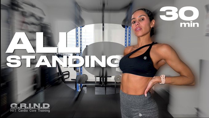 The image features a woman posing with her arms crossed while wearing a black sports bra and gray leggings. The background of the image shows a gym setting, indicating that she might be participating in an exercise challenge or fitness program. The caption on the image reads "All Standing" and there is also text below it stating "30 min All Standing".