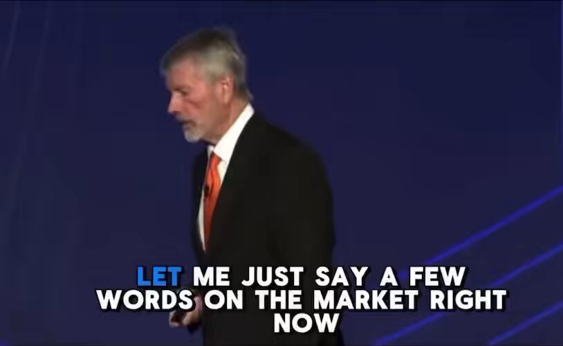 The image features a man wearing a suit and tie, standing against a blue background with white text that reads "Let me just say a few words on the market right now." The man appears to be speaking at an event or conference related to cryptocurrency or blockchain technology.