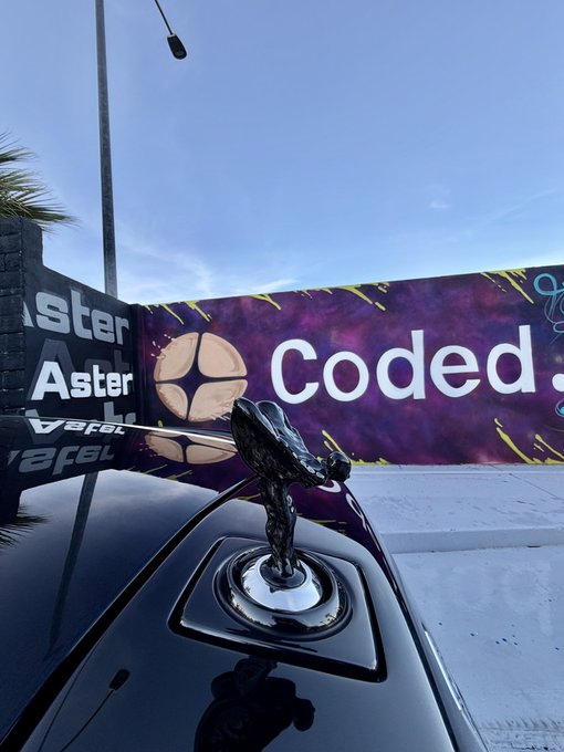 A sleek black Rolls-Royce hood ornament gleams in the foreground, with "Aster" and "Coded" branding visible on banners behind it against a twilight sky, tying into the post's promotion of the @Aster_DEX project.