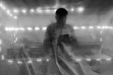 The image shows a man standing behind a sheet of plastic, possibly a shower curtain or a piece of fabric with holes cut out for air circulation. The photo appears to be in black and white, giving it an artistic and somewhat mysterious feel. The background is blurred, drawing attention to the man and his surroundings. The post reads "Life in Grayscale 0.235eth", which could be interpreted as a reference to the monochromatic nature of the image or possibly a description of the photo's subject matter.