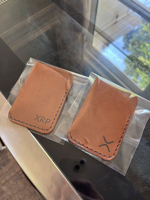 Two tan leather card sleeves in clear plastic packaging sit on a glass table, one embossed with "XRP" and the other with the XRP logo, ready to be shipped out to fans of the cryptocurrency.
