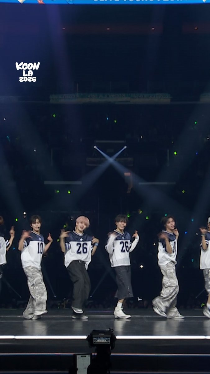 A group of performers dance on stage at KCON LA 2026, wearing matching white jerseys with the numbers "10," "26," and "26" visible, while the "KCON LA 2026" logo appears in the upper left and light sticks glow throughout the arena.